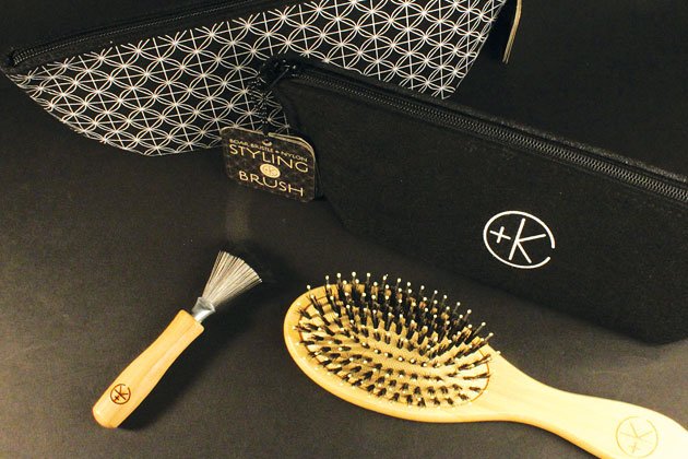 Two black zippered pouches, one bearing a geometric pattern and the other a plus-K logo, alongside a small brush and a large Cult and King Boar Bristle & Nylon Styling Brush | Brush Handle is 100% Bamboo from well-managed forests with neatly arranged bristles, all resting on a dark surface.