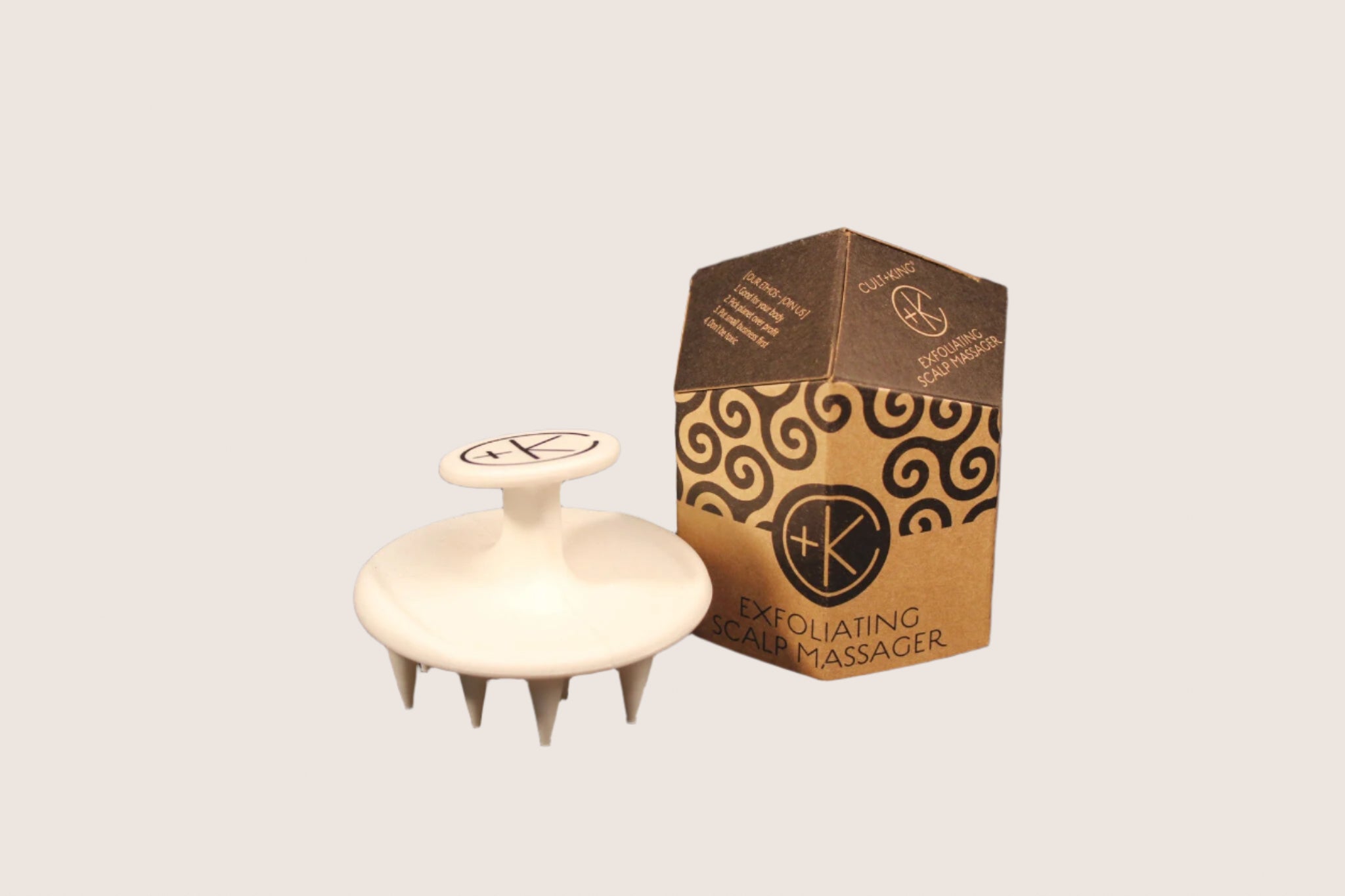 The **Cult and King Exfoliating Scalp Massager** from **Cult and King**, featuring white silicone with pointed bristles, is positioned beside its brown and black hexagonal packaging on a dark background.