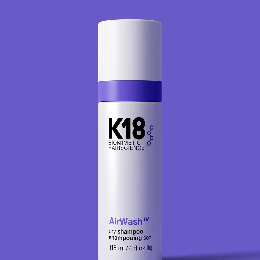 A white and purple bottle of K18 AirWash Dry Shampoo by Simply Colour Hair Salon Studio & Online Store, utilizing odorBIND biotechnology to reduce oil, containing 118 ml (4 fl oz). The bottle stands against a solid purple background.