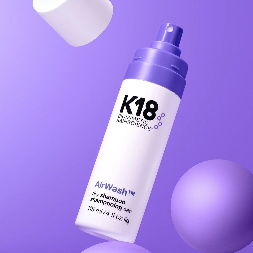 A bottle of K18 AirWash Dry Shampoo by Simply Colour Hair Salon Studio & Online Store, 118 ml, with the cap off, floats among purple spheres on a purple background. Utilizing odorBIND biotechnology, this product reduces oil and refreshes your hair effortlessly.