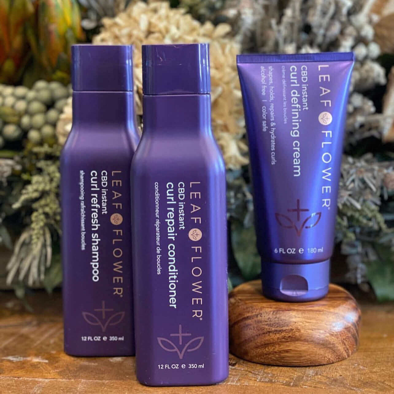 Three Leaf and Flower hair care products, including a Curl Refresh Shampoo, Leaf and Flower Instant Curl Repair Conditioner, and Curl Defining Cream, are displayed on a wooden surface with foliage in the background, ideal for all curl types and designed for optimal hydration and frizz reduction.