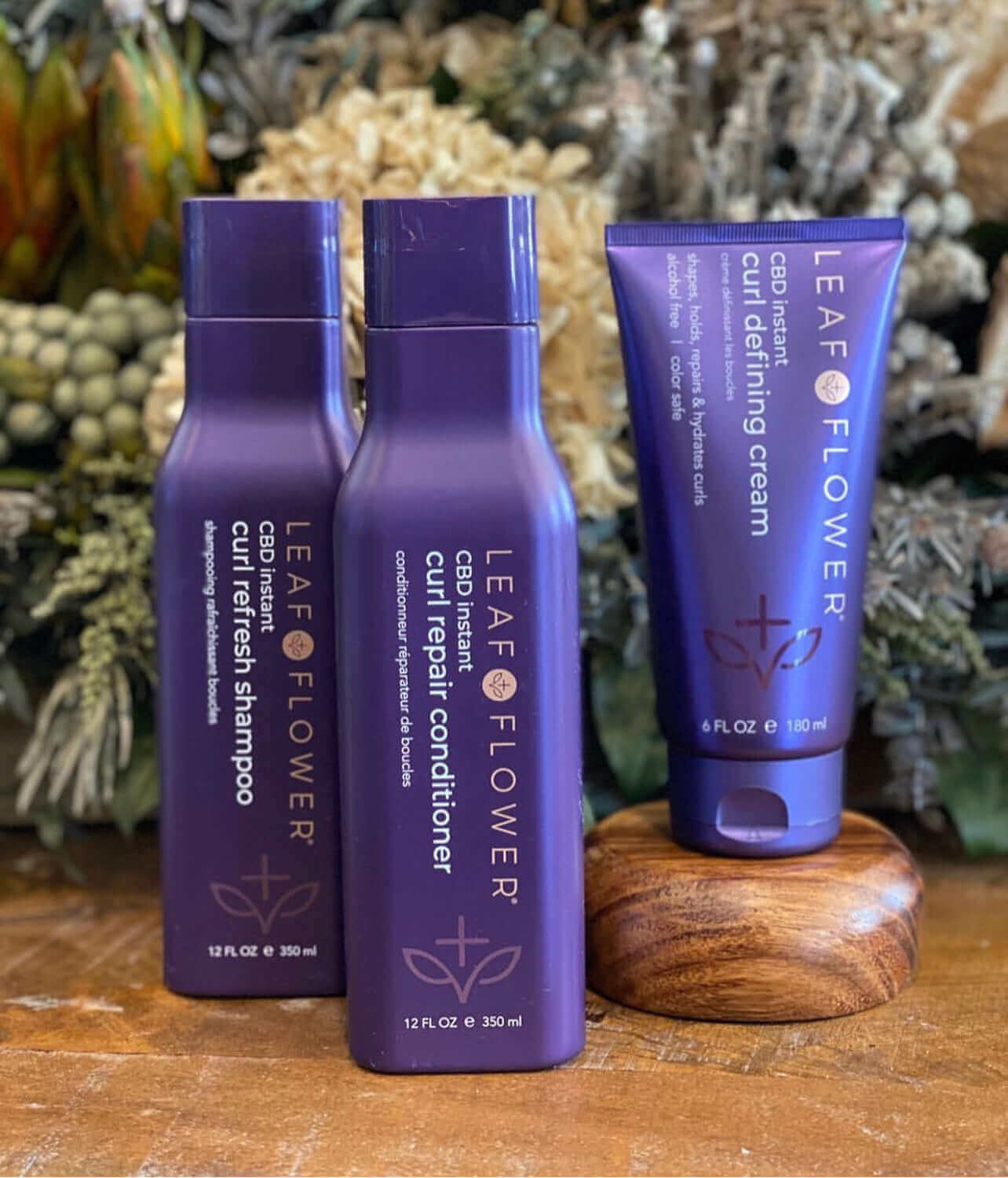 Three Leaf and Flower hair care products, including a Curl Refresh Shampoo, Leaf and Flower Instant Curl Repair Conditioner, and Curl Defining Cream, are displayed on a wooden surface with foliage in the background, ideal for all curl types and designed for optimal hydration and frizz reduction.