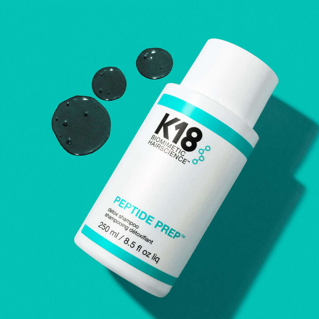 A white bottle of K18 Hair Repair K18 PEPTIDE PREP Detox Shampoo - Multiple Sizes lies on a turquoise background. The 250 ml (8.5 fl oz) bottle is complemented by some of its spilled contents, promising a deep cleanse with the power of K18PEPTIDE™ technology.