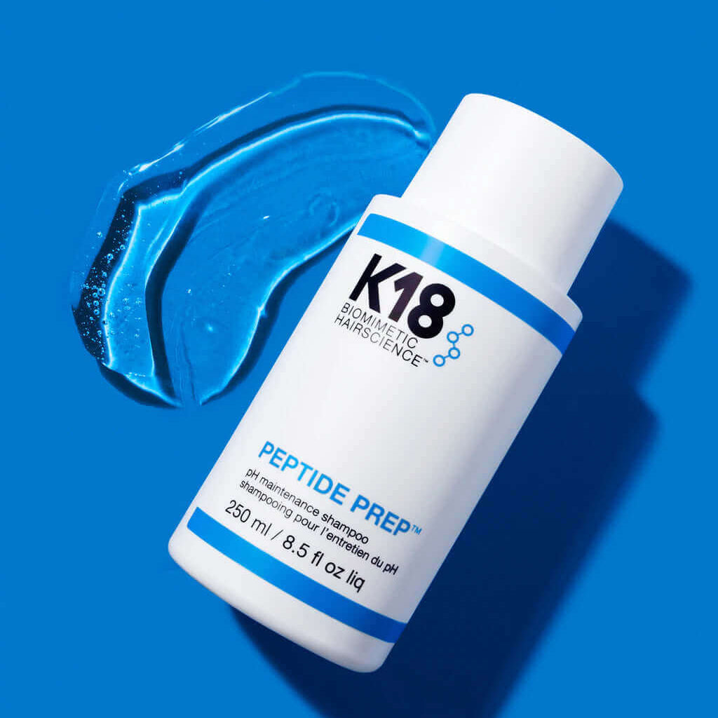 White bottle of K18 DAMAGE SHIELD pH Protective Shampoo - Multiple Sizes by K18 Hair Repair on a blue background, with a smear of the gel-like product beside it. Suitable for daily use to revitalize your hair, powered by the K18PEPTIDE™ formula.