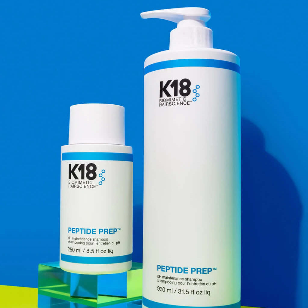 Two white bottles of K18 DAMAGE SHIELD pH Protective Shampoo - Multiple Sizes by K18 Hair Repair are displayed against a blue background. One bottle is 250 ml and the other is 930 ml, both labeled with volume and product name. Ideal for daily use, this cleansing shampoo ensures your hair stays fresh and vibrant.